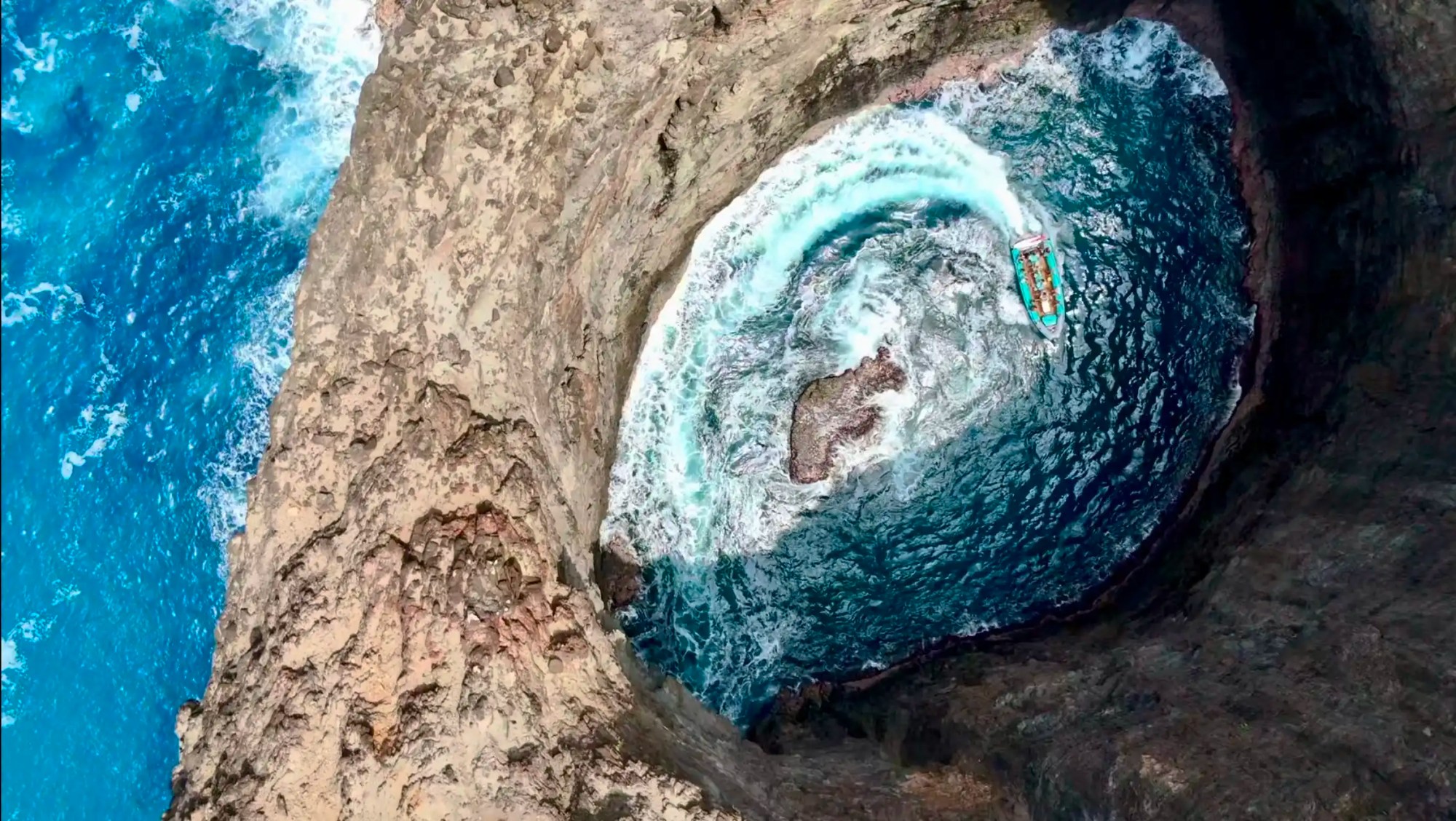 Aerial view of a boat in a circular sea cave with rough waves and rocky cliffs.