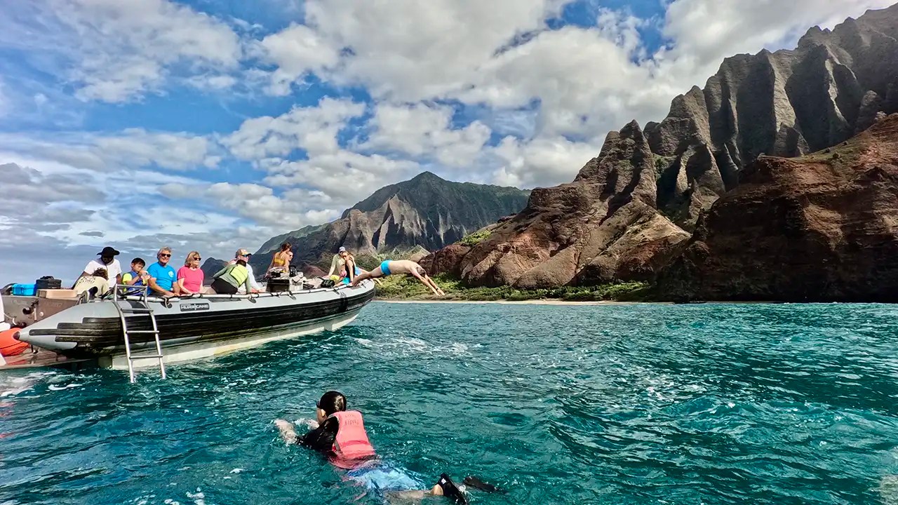 People on a boat, with one person diving, near cliffs under a blue sky.