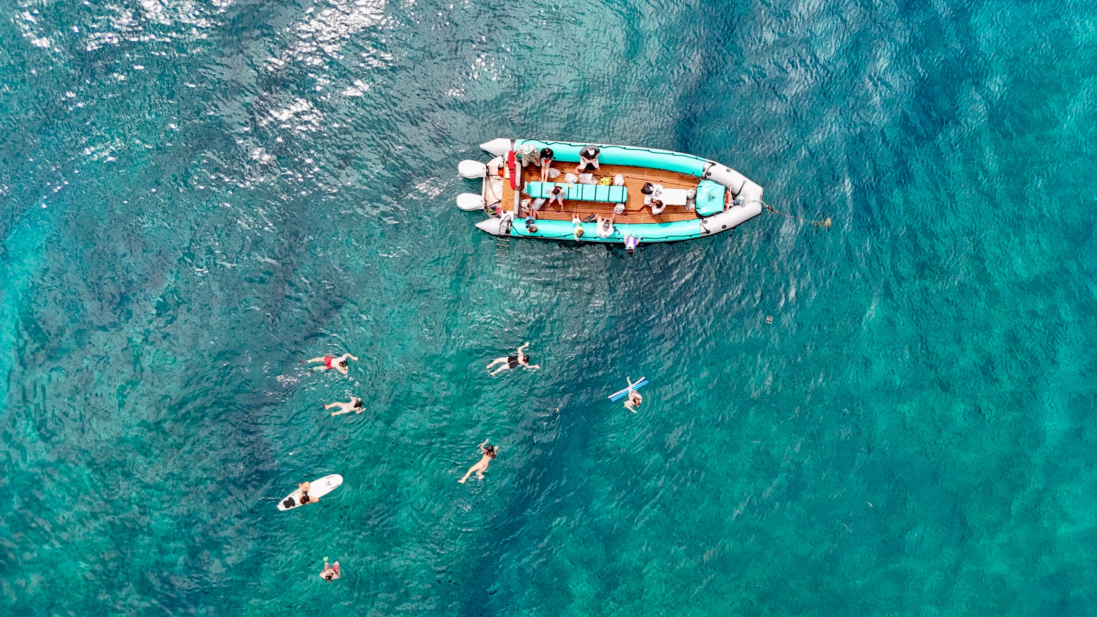 Aerial view of a boat with people, surrounded by swimmers in clear blue water.