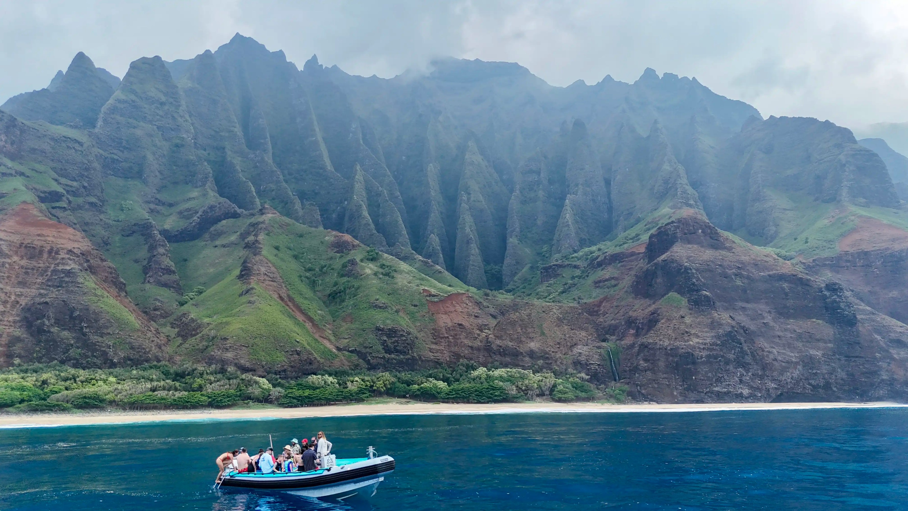 Boat with people on blue water near lush, rugged cliffs and cloudy sky.