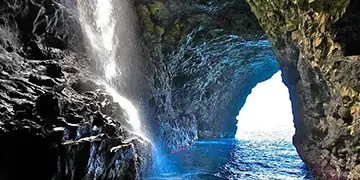 Sea cave with waterfall and blue water, sunlight entering through opening.