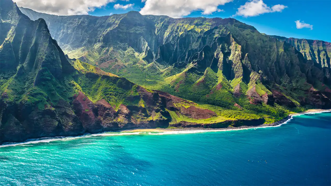 Lush green mountains with jagged cliffs beside a turquoise ocean under a blue sky.