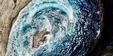 Aerial view of a boat making a circular wave pattern in the ocean near a rock.