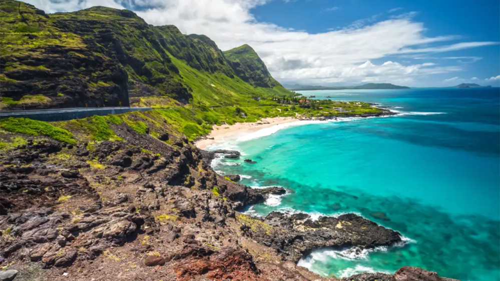 Coastal landscape with green cliffs, sandy beach, and turquoise ocean water.